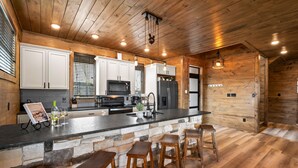 A modern kitchen with wooden cabinets, a large island with bar stools, a stone accent wall, and a hanging light fixture.