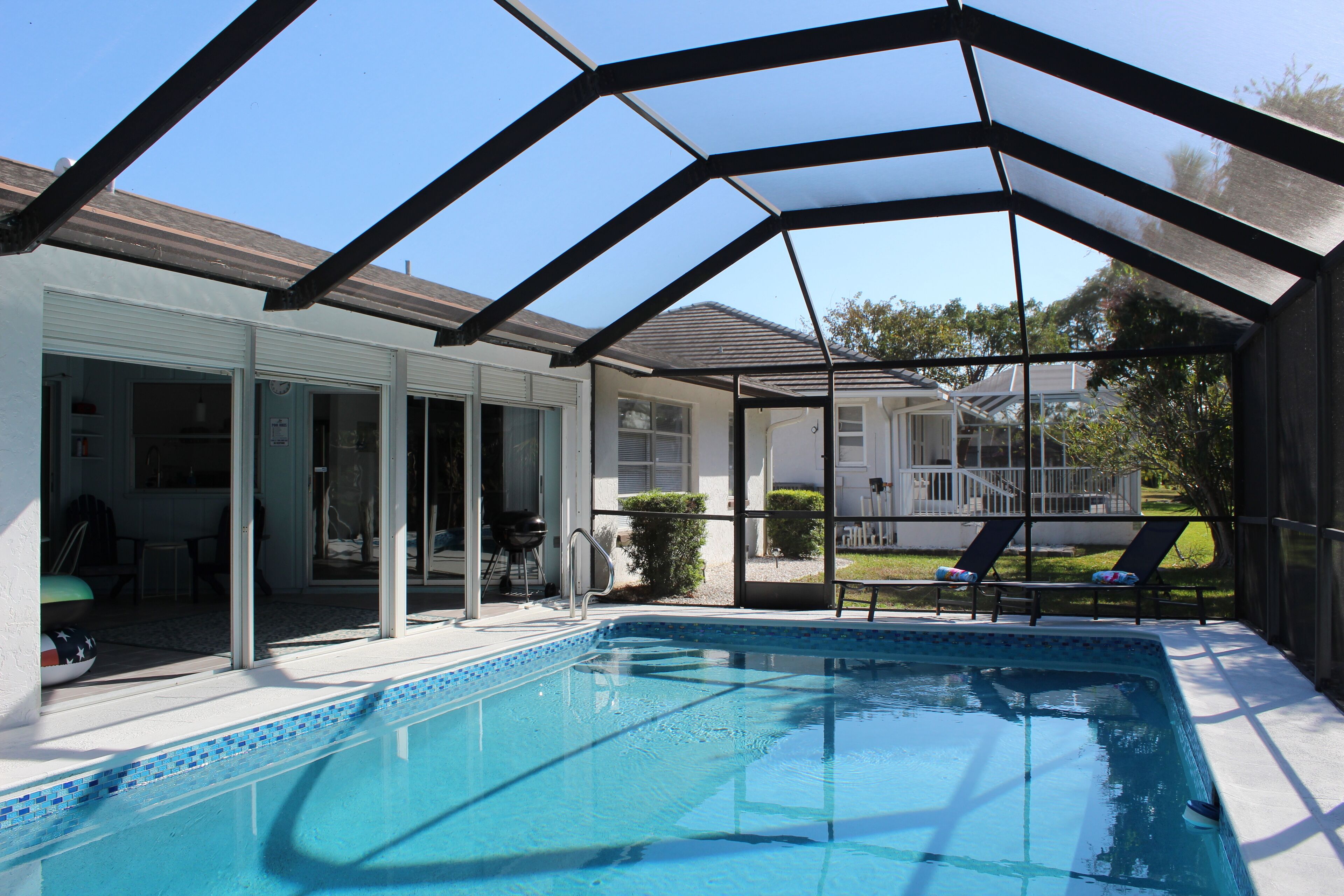 An indoor swimming pool with a glass enclosure, adjacent to a covered patio area with outdoor seating and a view of a residential neighborhood.