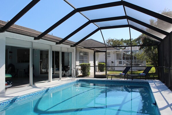 An indoor swimming pool with a glass enclosure, adjacent to a covered patio area with outdoor seating and a view of a residential neighborhood.