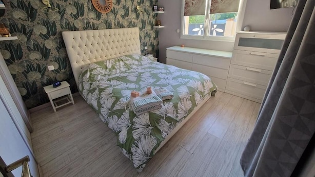Een slaapkamer met een bed, nachtkastje, raam en een televisie aan de muur.