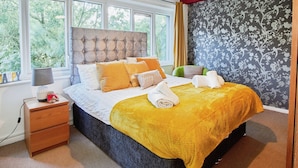 A bedroom with a large bed, a nightstand, a lamp, and a view of trees through the windows. - Charming City 1-bed Apartment in Birmingham (Birmingham)