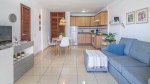 Un salon moderne avec un canapé gris, un coin repas avec une table et des chaises blanches, et une cuisine avec des meubles en bois et un réfrigérateur.