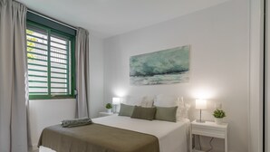 A bedroom with a bed, two bedside tables, a window with curtains, a painting on the wall, and a potted plant on the bedside table. - GO2TENERIFE Apart'Jara Pool view & Beach at 5 min (Playa de las Americas)