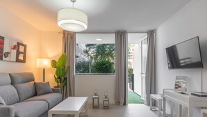 A modern living room with a grey sofa, a white coffee table, a TV, and a view of greenery outside. - GO2TENERIFE Apart'Jara Pool view & Beach at 5 min (Playa de las Americas)