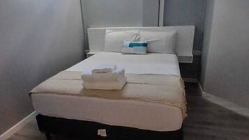 A small, compact bedroom with a single bed, a bedside table, a nightstand, a window, and a wall-mounted air conditioning unit.
