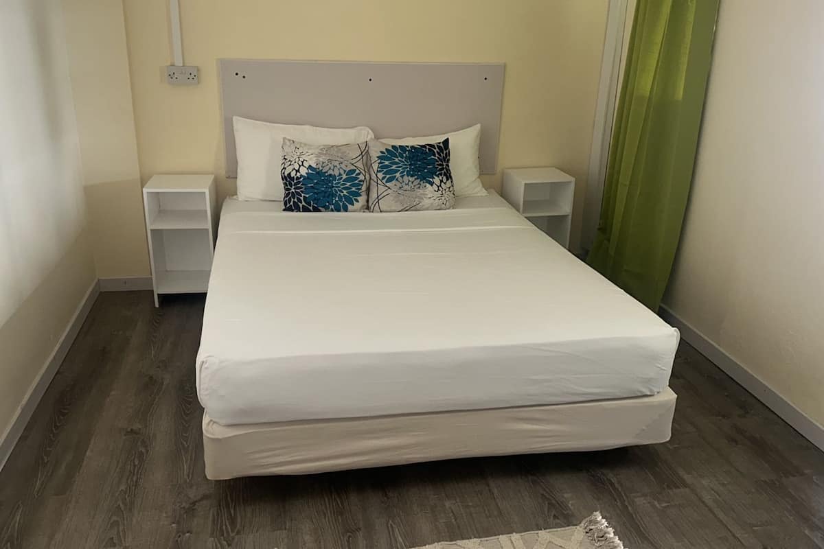Una cama bien hecha con ropa blanca y dos almohadones decorativos, flanqueada por mesitas de noche blancas, en una habitación con piso de madera y un diseño sencillo y limpio.