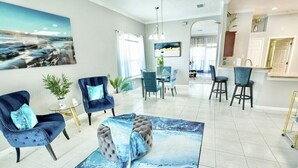 Een moderne woonkamer met een vloerkleed met marmerprint, blauwe fauteuils, een rond hockertje, en een eethoek met een glazen tafel en stoelen.