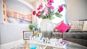 Een woonkamer met een glazen salontafel, een vaas met roze bloemen, een bord met daarop 'welkom' en een schilderij van een stadsgezicht.