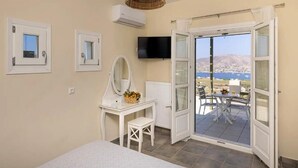 Ein Schlafzimmer mit einem Bett, einem Schminktisch mit Spiegel und einer Kommode. An der Wand ist ein Fernseher und es gibt ein Fenster mit Blick auf das Meer.