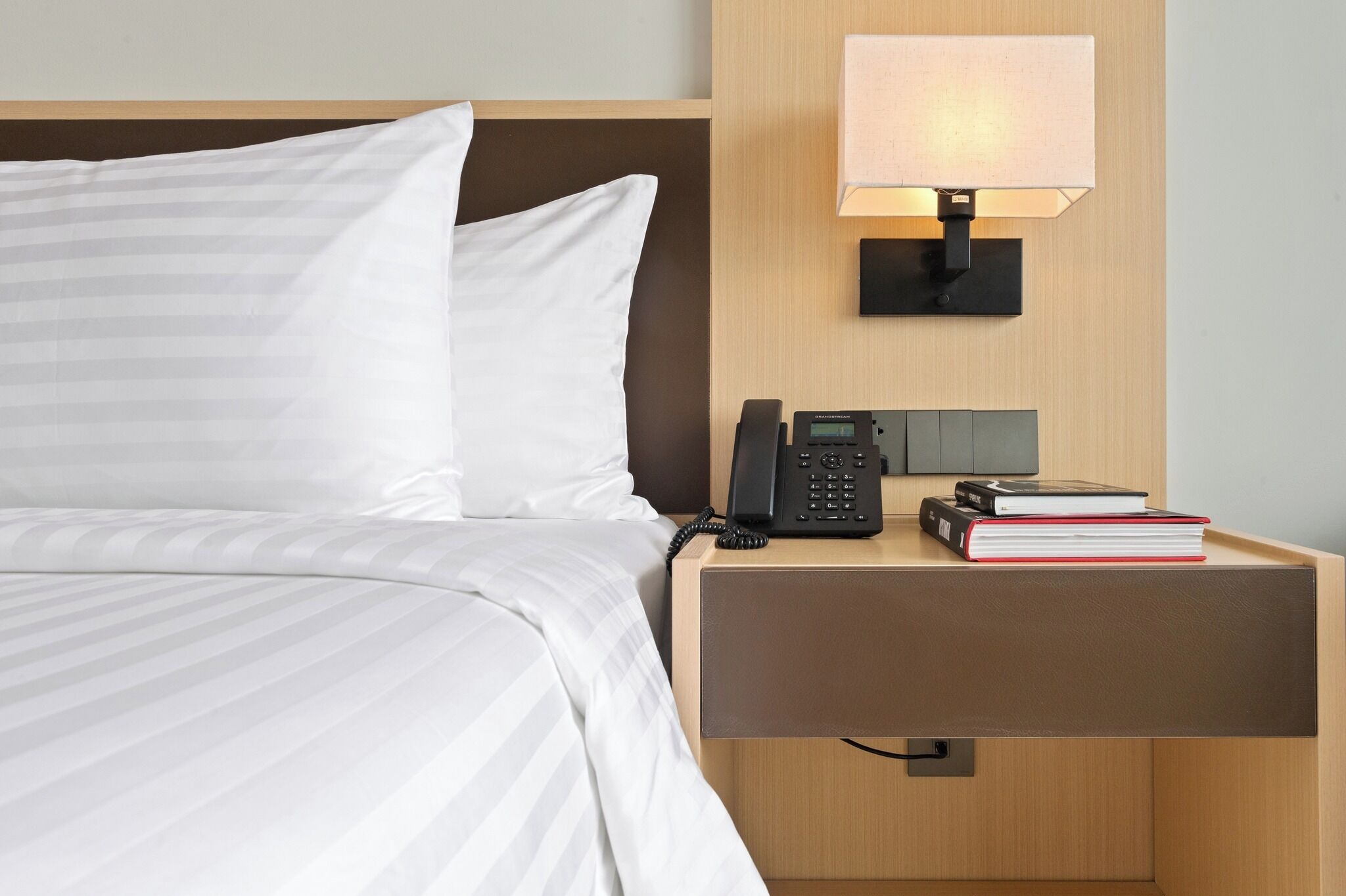 A neatly made bed with white bedding, a bedside table with a telephone, a lamp, and a few books.