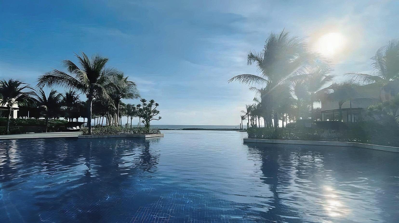 A clear blue infinity pool with palm trees on both sides, a view of the ocean, and a bright sun in the sky.