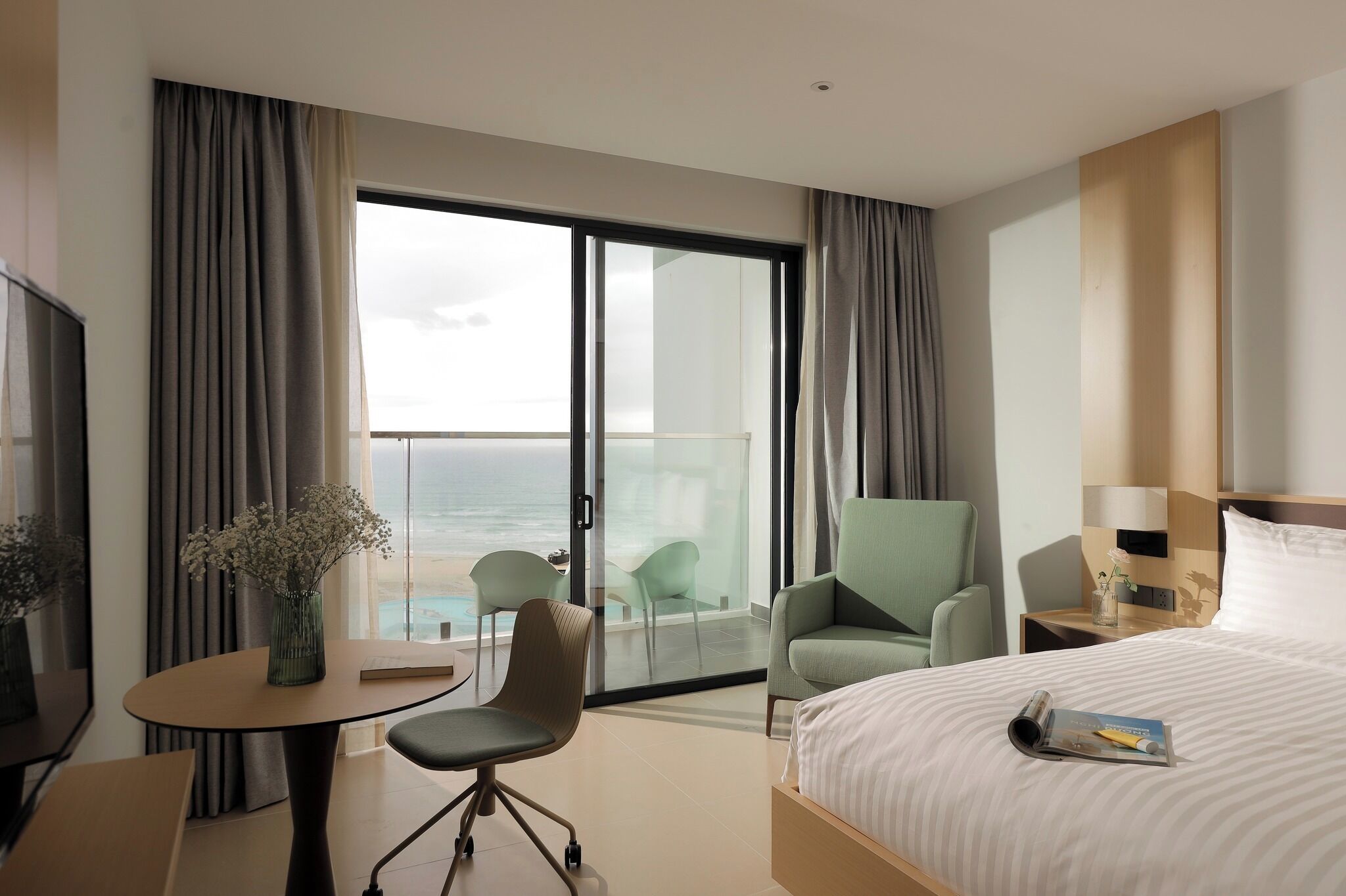 Une chambre d’hôtel avec un lit, un bureau et une chaise, une petite table avec un vase, et une vue sur l’océan par une porte-fenêtre coulissante.