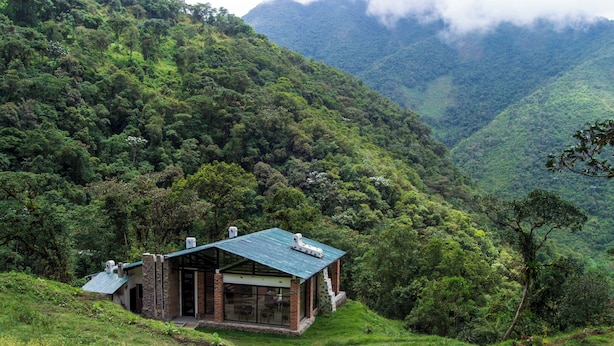 Una casa de montaña apartada con techo verde, rodeada de abundante vegetación y montañas.