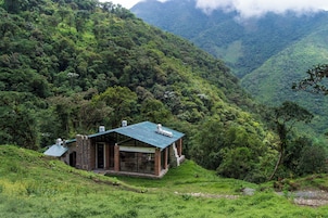 Una casa de montaña apartada con techo verde, rodeada de abundante vegetación y montañas.
