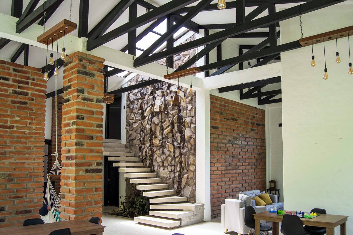 Interior moderno com paredes de tijolo aparente, uma escadaria e um espaço de refeições com mesa e cadeiras.
