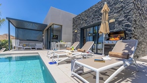 Un área de piscina moderna al aire libre con sillas de descanso, una parrilla y un muro de piedra.