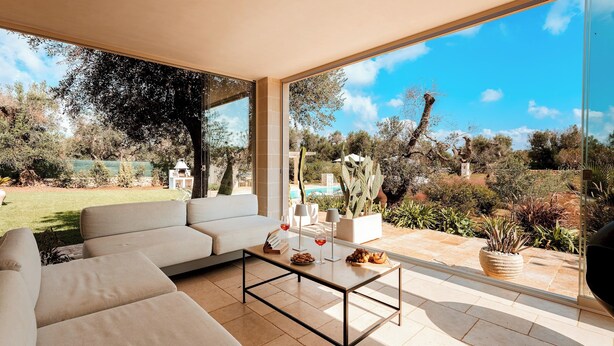 A spacious living area with a large window, a beige sofa, a coffee table with snacks and drinks, and a view of a garden and pool. - Villa Belkis 6, Emma Villas (Ostuni)