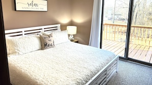 A bedroom with a bed, a bedside table with a lamp, a window with a view of a deck, and a wall sign. - Life-Giving, Quiet retreat only 25 min west of Indpls. Peace, Pace, & Gratitude (Coatesville)