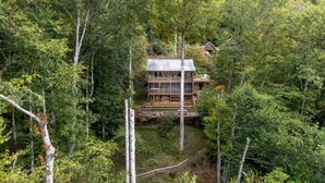 Una cabaña rodeada de un bosque frondoso, con una terraza visible y un amplio ventanal en la planta principal.