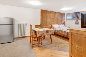 Quarto compacto com cama, mesa com cadeiras, armário de madeira, radiador e frigorífico.