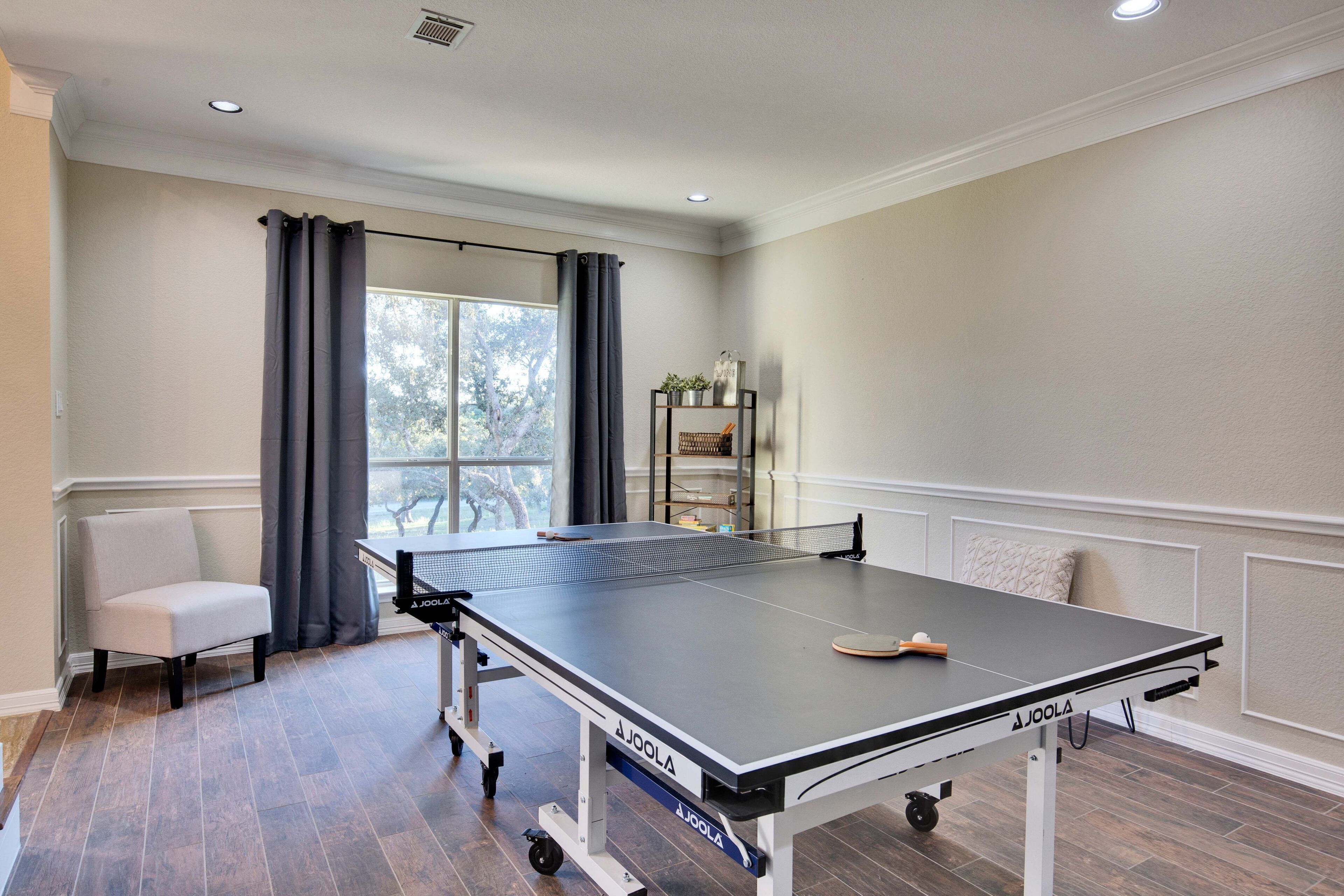 Une table de ping-pong dans une pièce avec une fenêtre, une chaise et une étagère remplie d’objets.