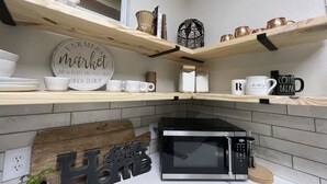 Een keuken met houten planken vol servies, een bord met de tekst 'Farmers Market Verse Groenten en Fruit Elke Dag Geopend', en een magnetron op het aanrecht.