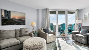 Un salon moderne avec un canapé, des fauteuils et un pouf à motifs. Un tableau représentant un coucher de soleil est accroché au mur, et de grandes portes vitrées offrent une vue sur le paysage urbain.