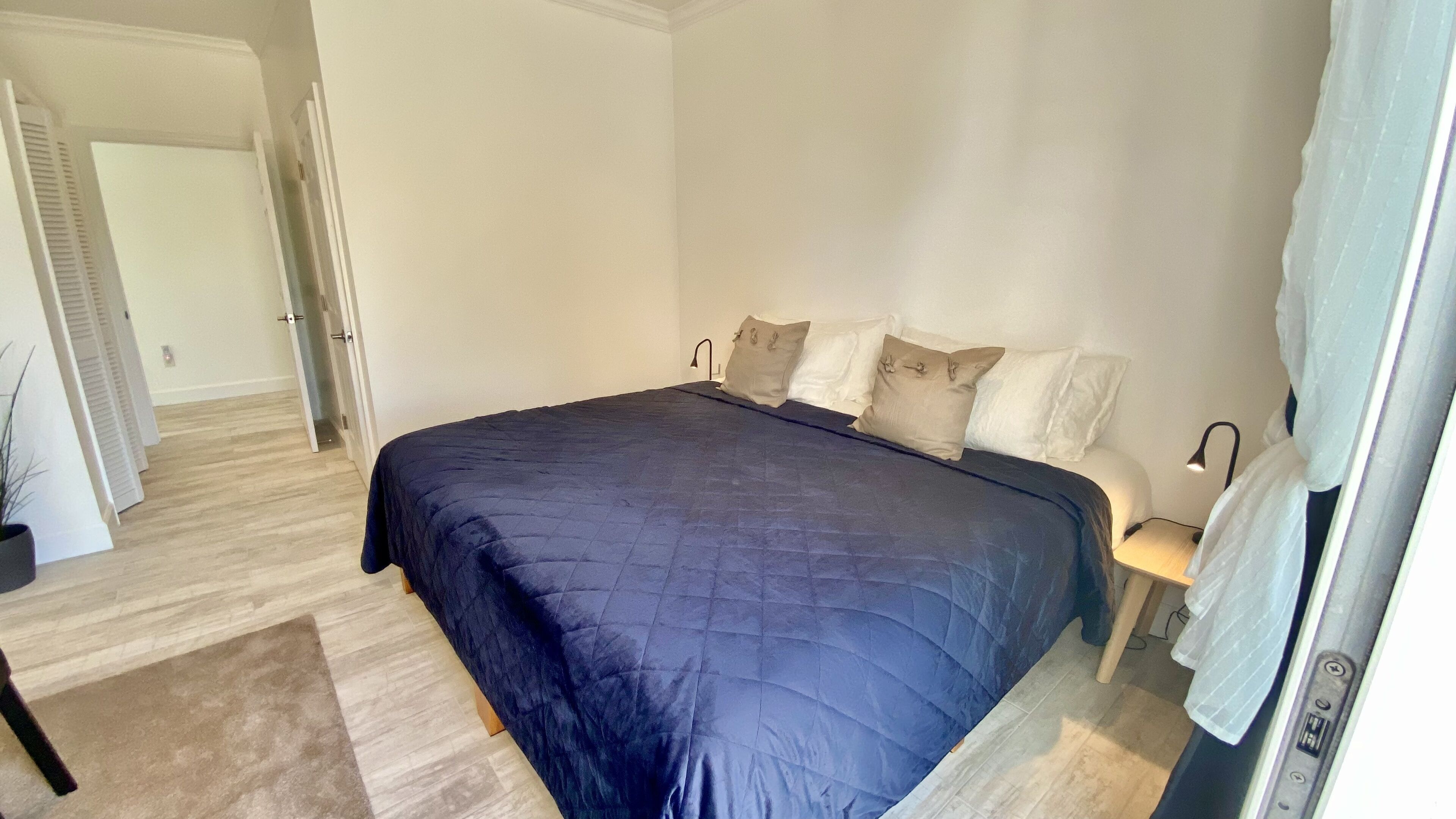 A bedroom with a large bed, a bedside table with a lamp, and a door leading to another room.