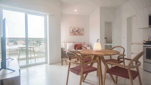A modern living room with a glass door leading to a balcony, a dining area with a round wooden table and wicker chairs, and a kitchen area with white cabinets.
