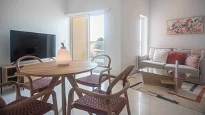 A modern living room with a round wooden table, wicker chairs, a flat-screen TV, a sofa with cushions, and a large window offering a view.