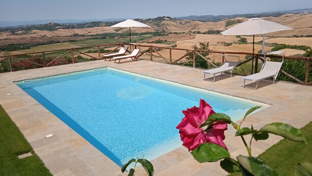 Une piscine rectangulaire à l’eau d’un bleu limpide, entourée d’une terrasse en pierre. Deux parasols blancs offrent de l’ombre, et plusieurs chaises longues sont disposées autour de la piscine. La piscine se trouve dans un cadre de collines verdoyantes et sous un ciel dégagé.