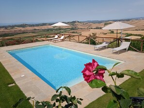 Ein rechteckiger Swimmingpool mit glasklar blauem Wasser, umgeben von einer Steindeck. Zwei weiße Sonnenschirme spenden Schatten, und mehrere Liegestühle sind rund um den Pool platziert. Im Hintergrund sind sanfte Hügel und ein klarer Himmel zu sehen.