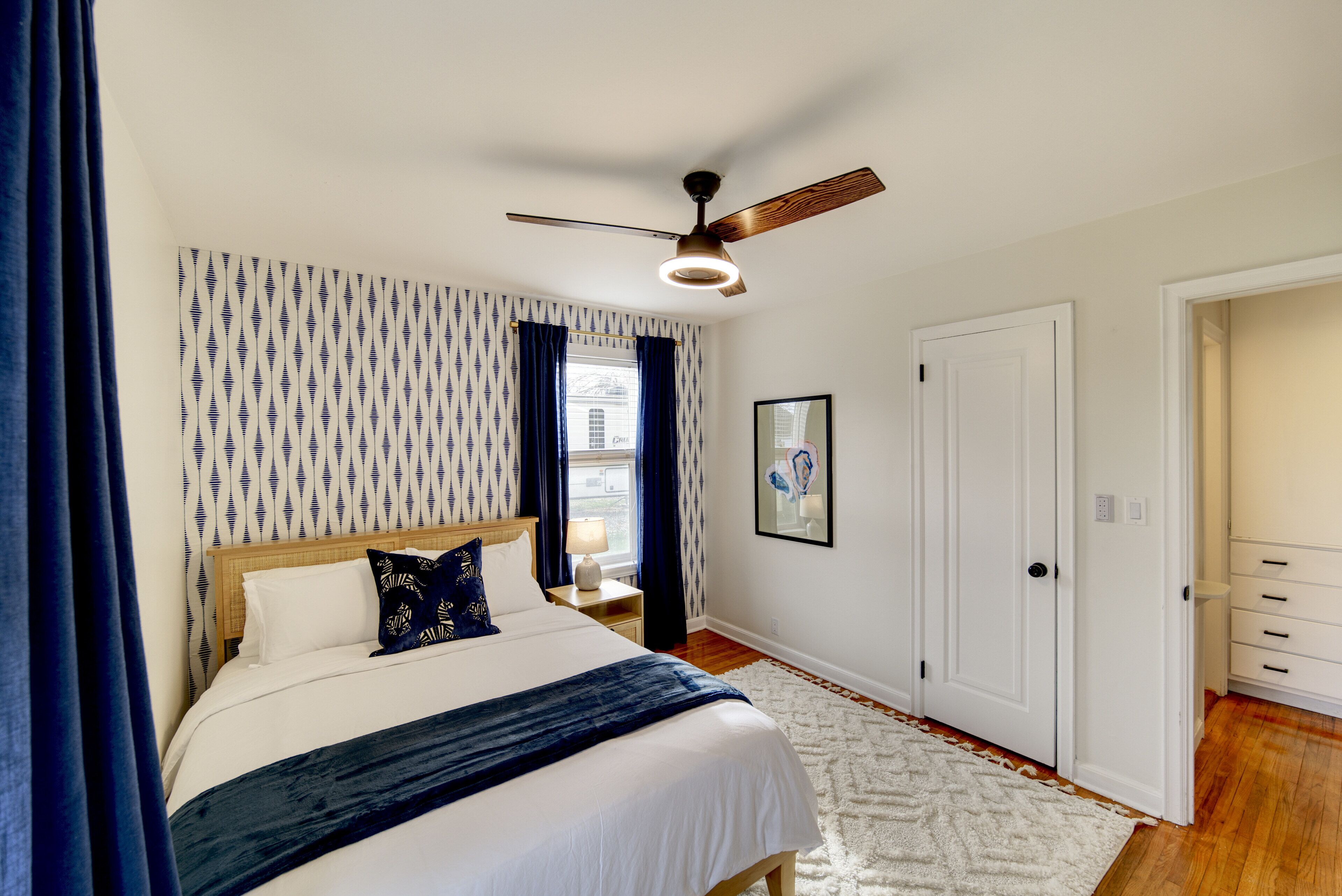 A bedroom with a bed, a ceiling fan, a nightstand, a white door, and a patterned wall.