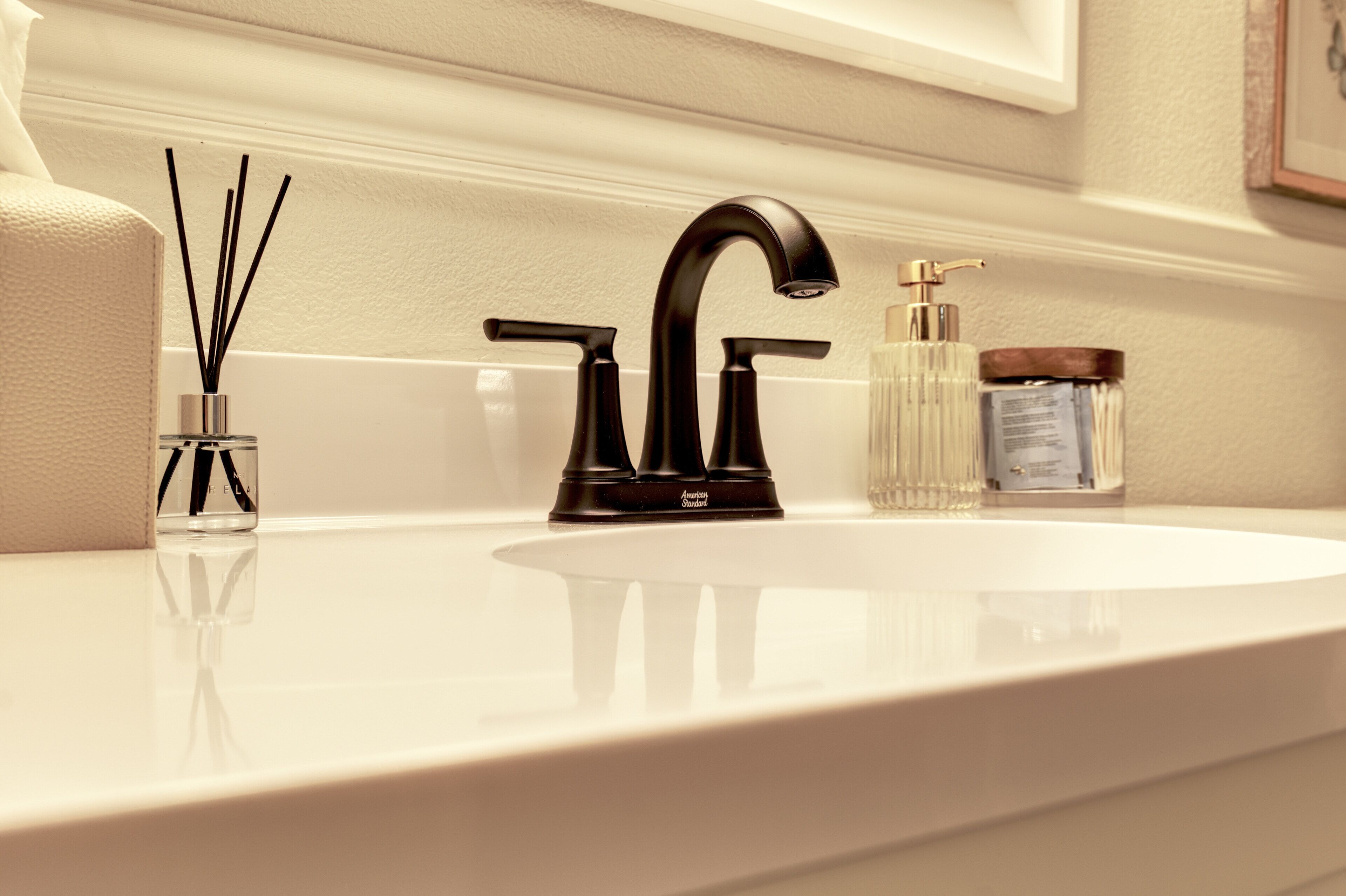 A bathroom sink with a black faucet, a clear glass container with black reeds, and a clear glass bottle with a pump.