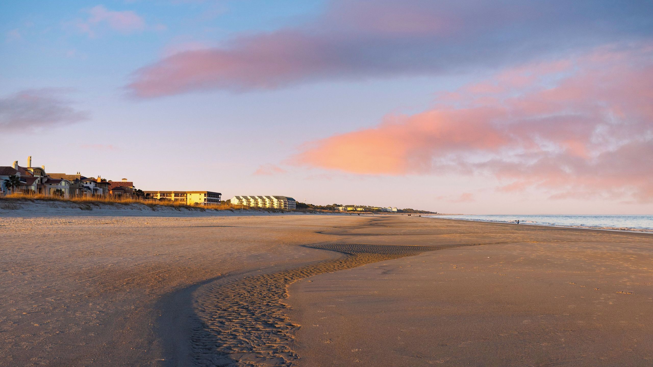 A beach with a curved sand pattern, buildings in the background, and a sunset sky.