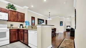 A kitchen with wooden cabinets, a white dishwasher, and a granite countertop. There is a dining area with a table and chairs, and a hallway leading to a bedroom. - Awesome Vacation Destination! 1304 Tuscana Resort (Davenport)