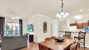 A spacious living room with a gray sofa, a wooden dining table with chairs, a TV mounted on the wall, and a kitchen area with wooden cabinets and a countertop. - Awesome Vacation Destination! 1304 Tuscana Resort (Davenport)