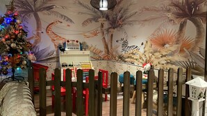 Una stanza con un murale raffigurante animali, un albero di Natale e un’area giochi con giochi e un pianoforte giocattolo.
