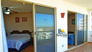 Un dormitorio con cama, una puerta corrediza de vidrio que da a un balcón, y el logotipo de Rocky Point Booking.
