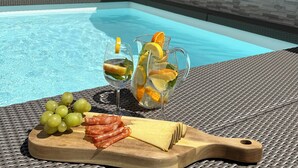 Ein Poolbereich mit Korbstuhl, Holzschneidebrett mit Trauben, Käse und Aufschnitt, sowie eine Limonadenkanne mit Fruchtscheiben.