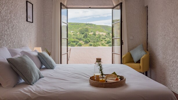 Ein Schlafzimmer mit Bett, Kissen, einem Nachttisch mit Lampe, einem Sessel und Blick auf eine grüne Landschaft durch eine geöffnete Tür.