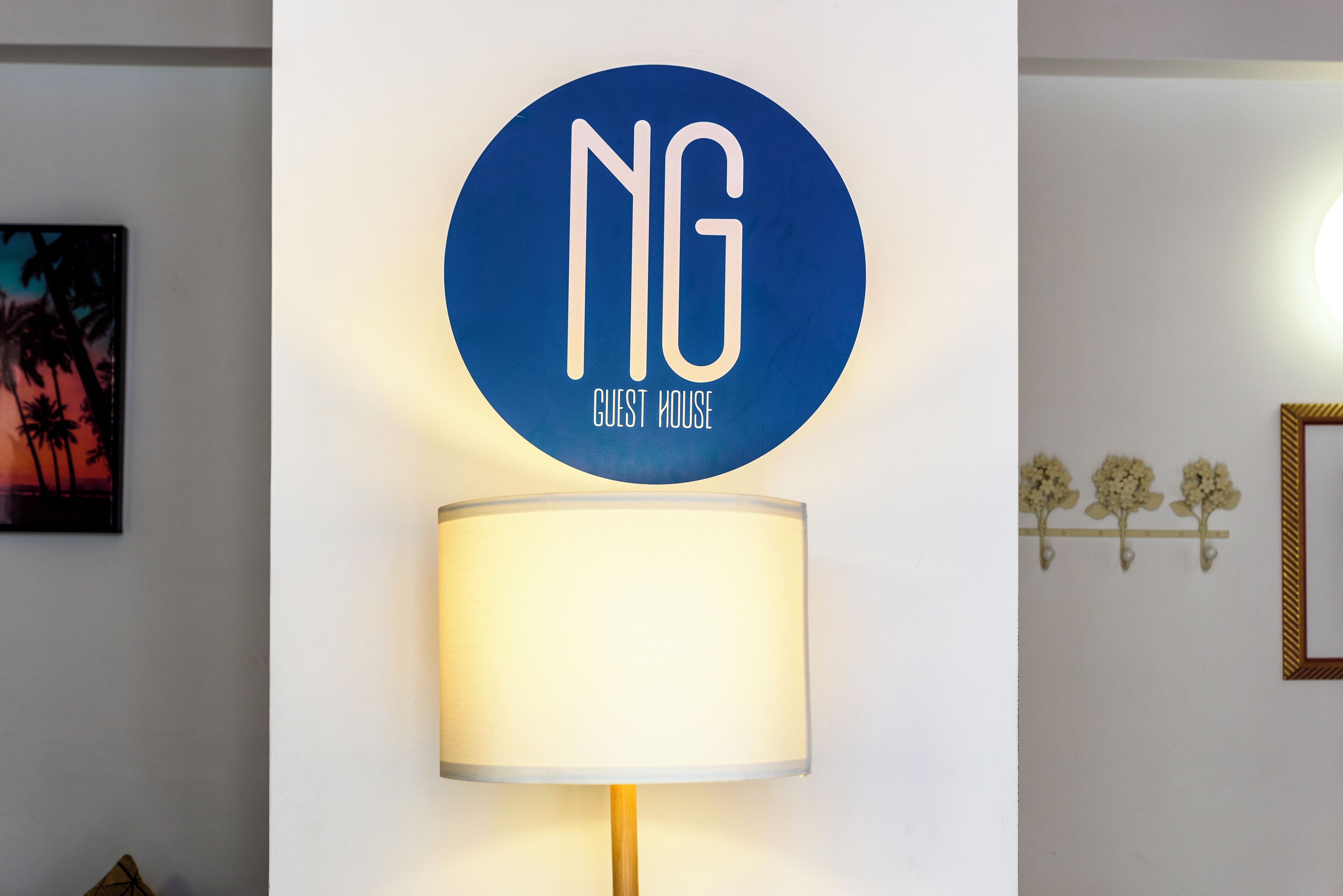 A wall-mounted sign with 'NG' and 'GUEST HOUSE' text, a wall-mounted lamp, and a framed picture with palm trees.