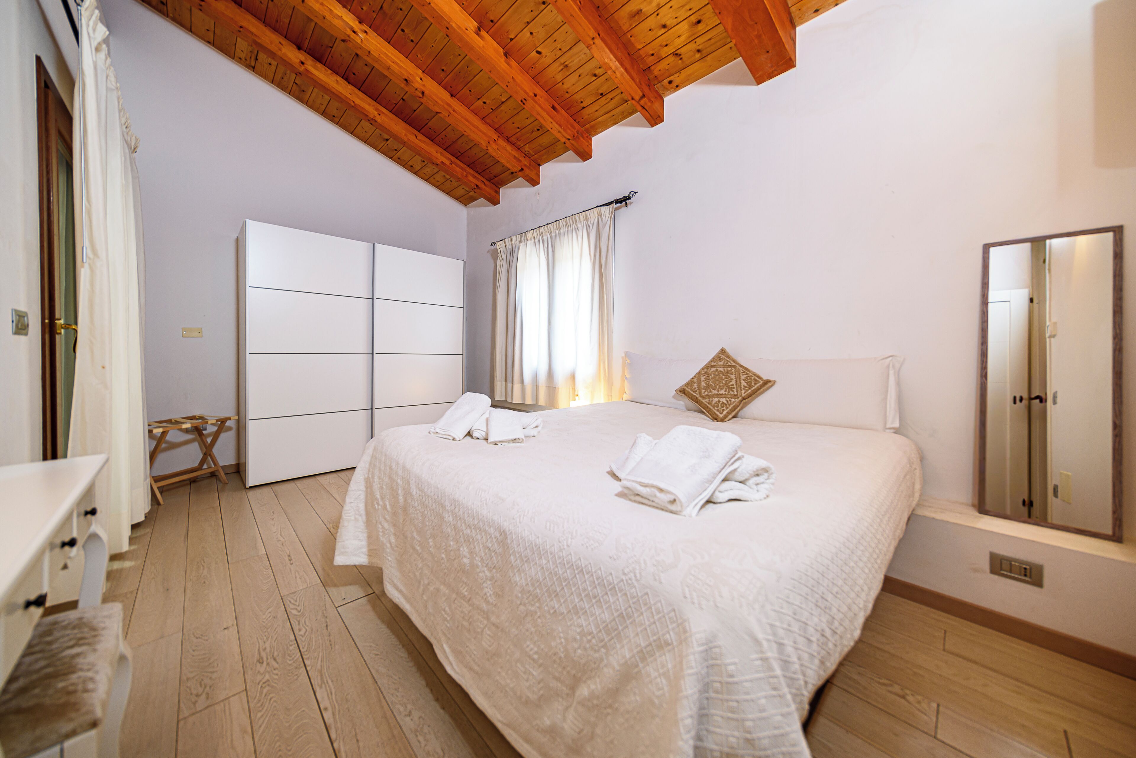 A bedroom with a wooden ceiling, a bed with white linens, a bedside table, a mirror, and a window with curtains.