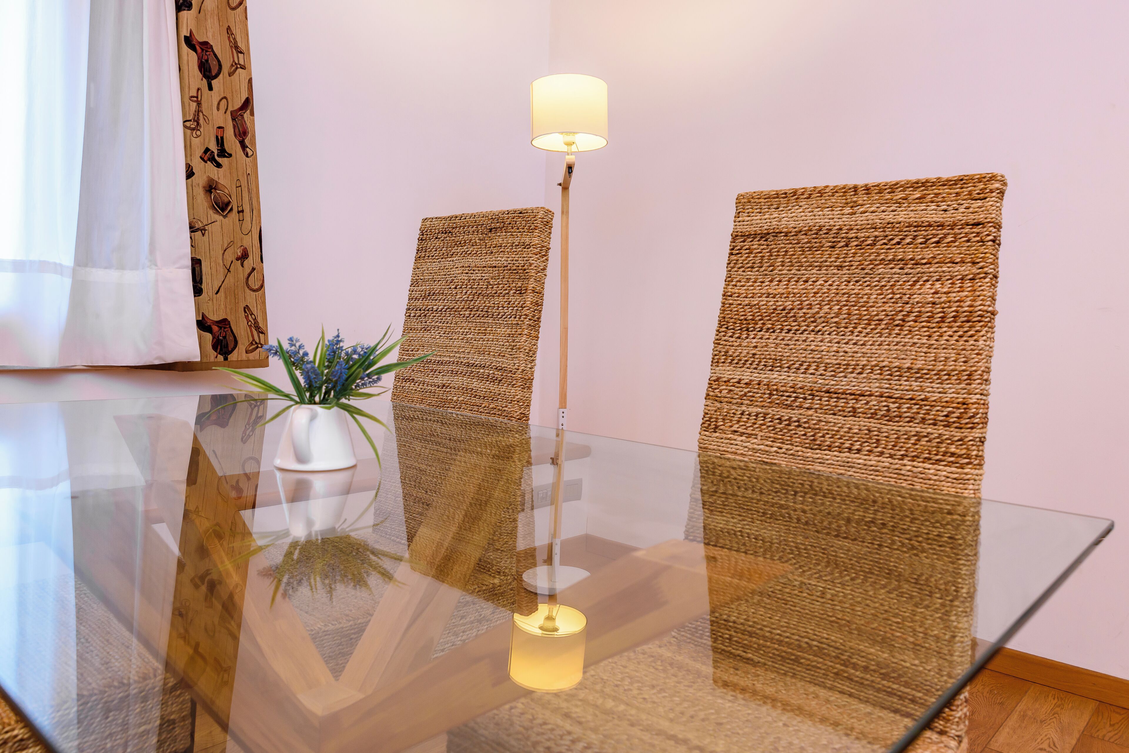 A glass-top table with a white vase and a plant, two woven backrests, and a floor lamp.