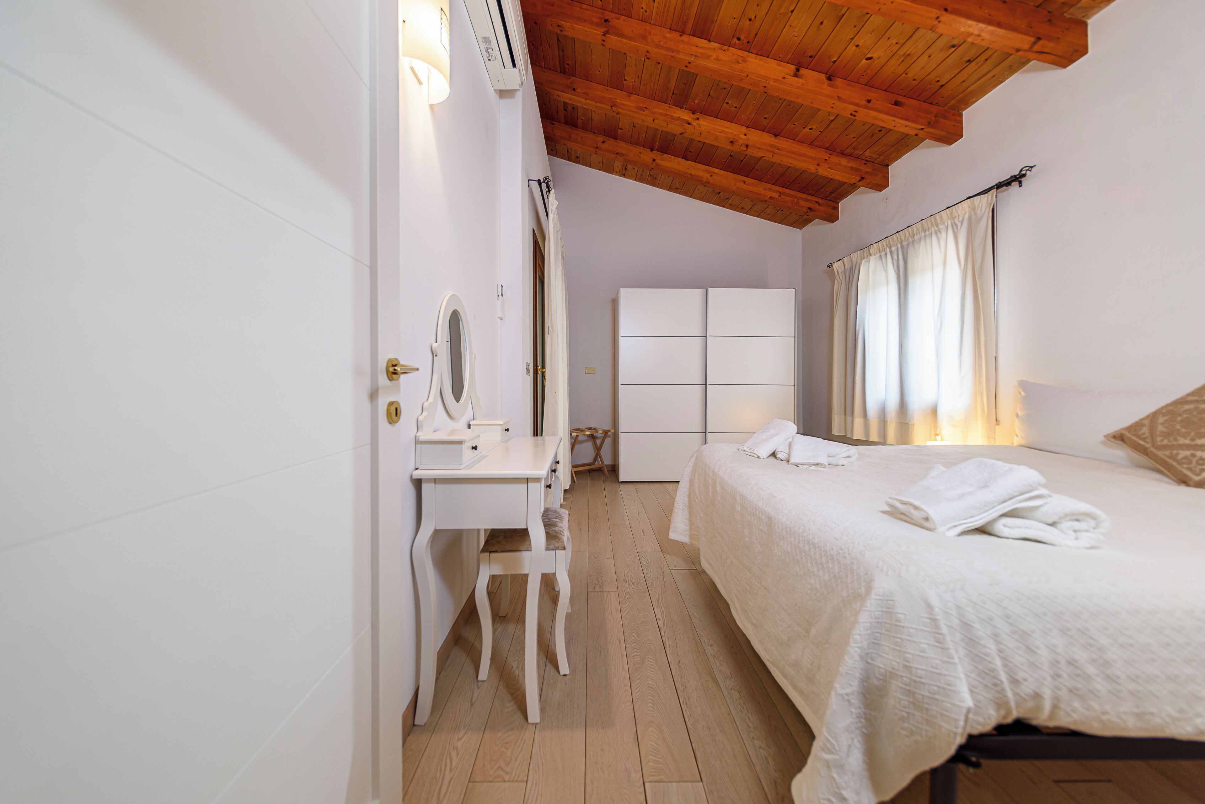 A bedroom with a wooden ceiling, a white desk with a mirror, a bed with white linens, and a wardrobe.