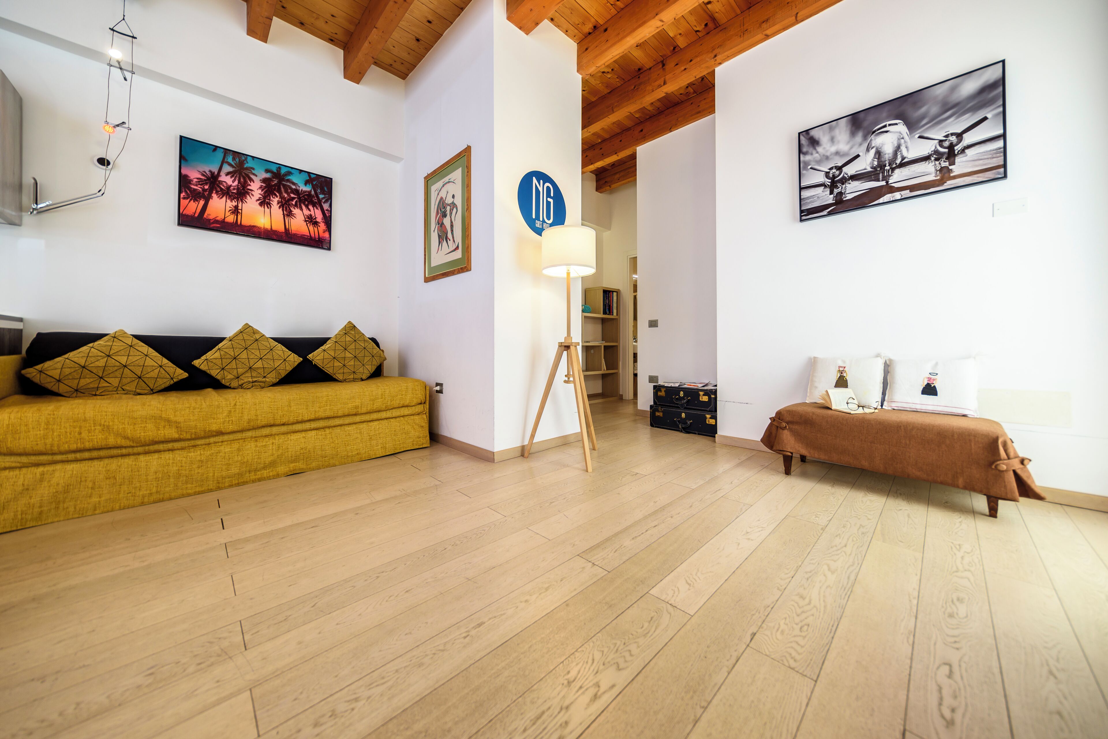 Une pièce avec un plancher en bois, un canapé jaune, un banc marron et deux tableaux accrochés aux murs.