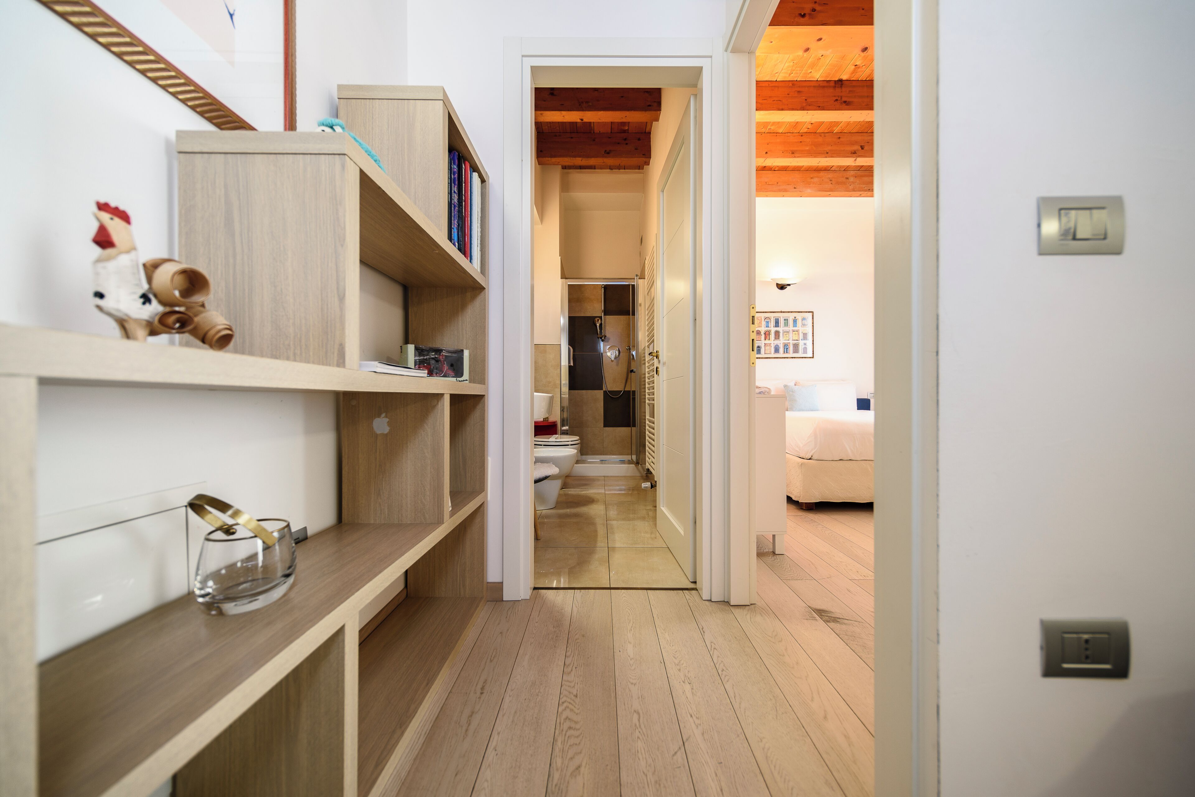 Un pasillo estrecho con suelo de madera, una pared blanca y una puerta que da a una habitación con cama y radiador.