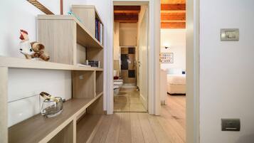 A narrow hallway with wooden flooring, a white wall, and a door leading to a room with a bed and a radiator.
