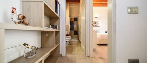 Un pasillo estrecho con suelo de madera, una pared blanca y una puerta que da a una habitación con cama y radiador.