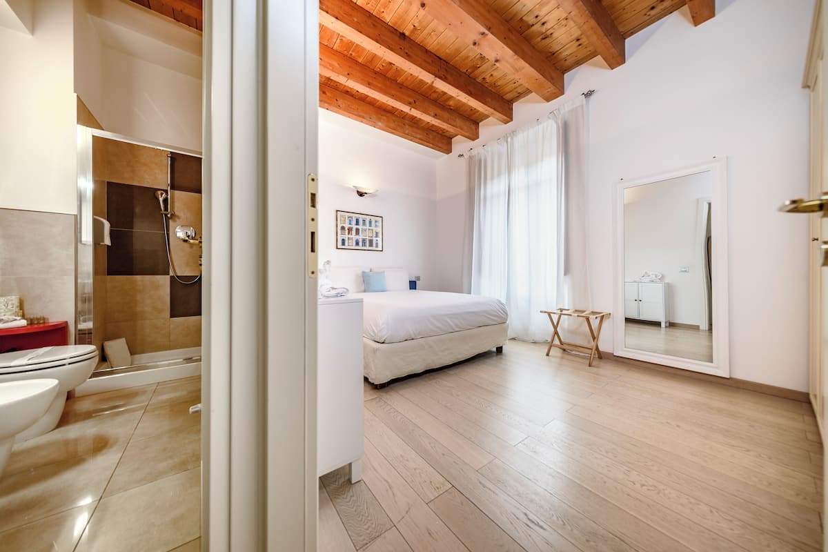 Quarto com cama, mesa de cabeceira, espelho, chuveiro e vaso sanitário.